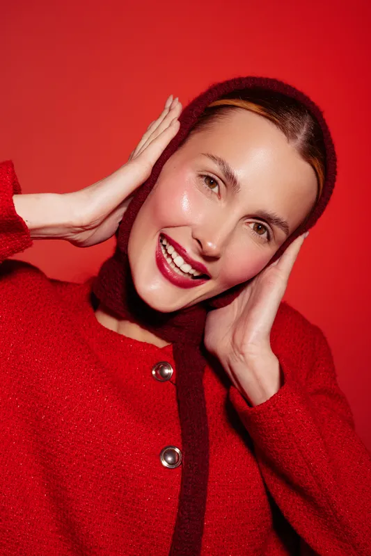Smiling woman in red sweater poses with hands near face against red background. Close-up portrait with natural makeup and joyful expression. Concept of happiness, fashion, and beauty.