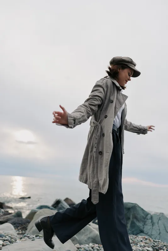 Woman in a long coat and hat balances on rocks by the sea during cloudy weather. Casual autumn outfit and natural outdoor activity near water. Concept of freedom and nature.