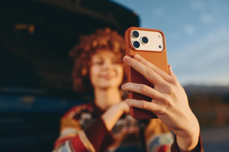 Woman takes selfie with smartphone outdoors, her hand holding the phone in front, car nearby, blurred portrait background, warm lifestyle mood, casual travel moment, sunlight and sky