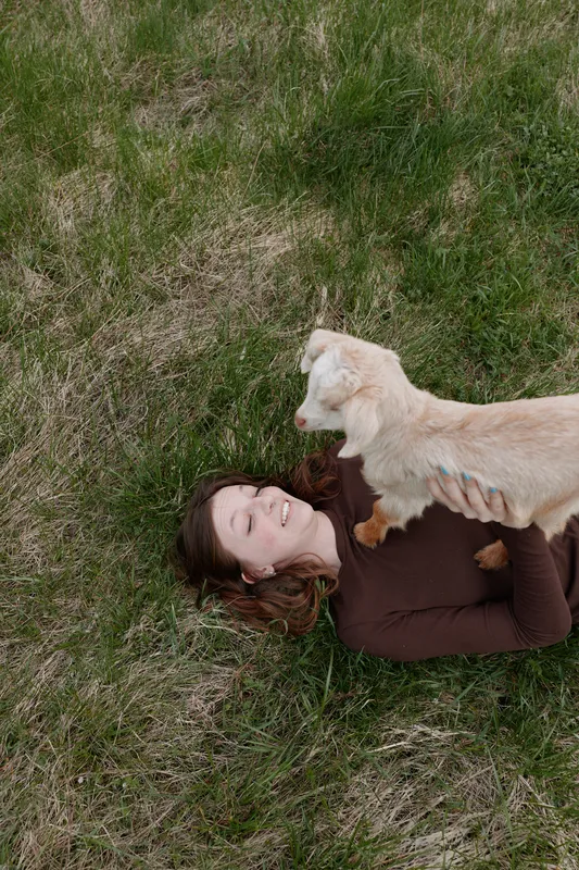 Goat kid resting on a smiling woman lying in grass, outdoors lifestyle moment, playful affection, sunlit meadow scene, natural colors, relaxed pose, gentle bond, carefree mood, rustic charm.