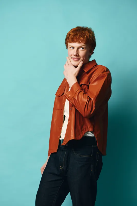 Young man with red hair in brown leather jacket posing with hand on chin against blue background. Casual style, confident expression and studio portrait with copy space.