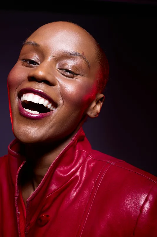Close-up of happy black woman with dark skin laughing, wearing red leather jacket on black background. Concept of joy, fashion, confidence, and beauty in portrait photography.