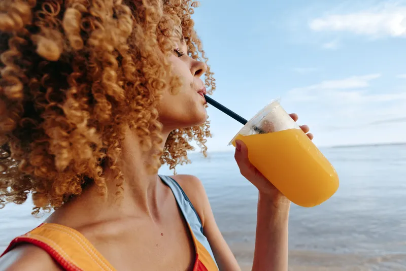 Young woman with curly hair drinks orange juice from a plastic cup with a straw while standing by the calm sea under a clear sky on a warm day.