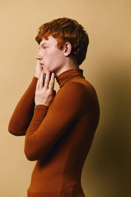 Young man with red hair in brown turtleneck standing in profile against beige background, thoughtful expression and hand near face in studio portrait.