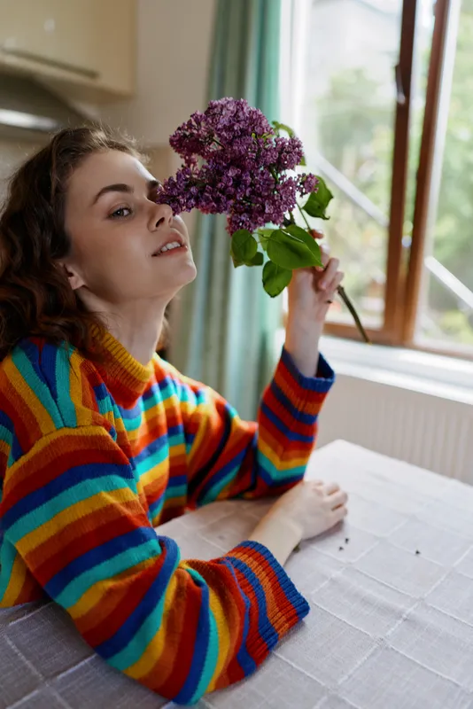 Young woman sits at a bright home table near a window, holding a purple bouquet, wearing a rainbow striped sweater, relaxed and smiling in a cozy lifestyle portrait