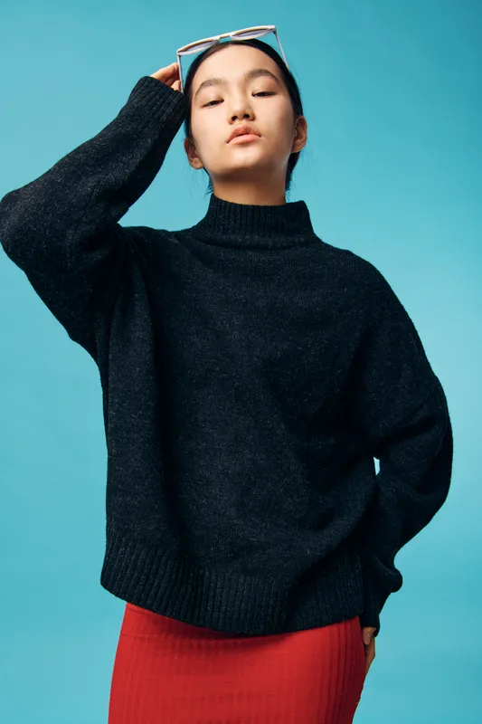 Young woman in black sweater and red skirt standing with hand on head against blue background. Fashion model posing with closed eyes and casual confident expression in studio.