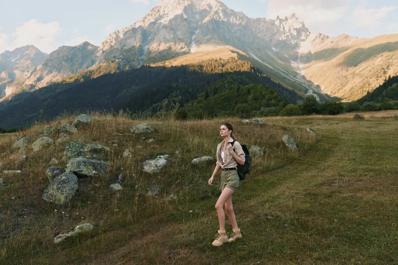 Woman hiker walking through a meadow beside rocky mountains, carrying a backpack, enjoying outdoor nature and expansive scenery in daylight