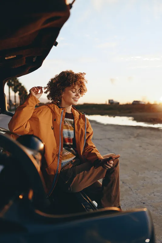 Woman lifestyle outdoors by the beach at sunset, curly haired person smiling while sitting near a car door, wearing a warm jacket, golden light, relaxed travel mood