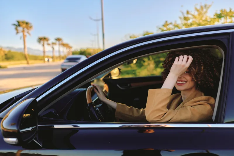 Cheerful woman driving outdoors, smiling with eyes closed, sunlit moment, relaxed posture, retro vibe, lifestyle and casual mood, colors warm and natural