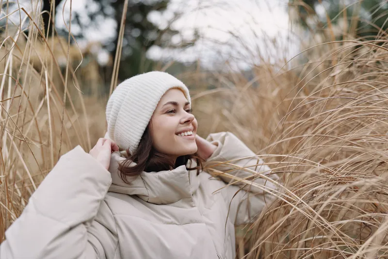 Woman enjoying a chilly outdoor moment in a beige coat and white beanie amidst tall grass, smiling softly, capturing cozy winter mood and nature's calm in a scenic field setting.