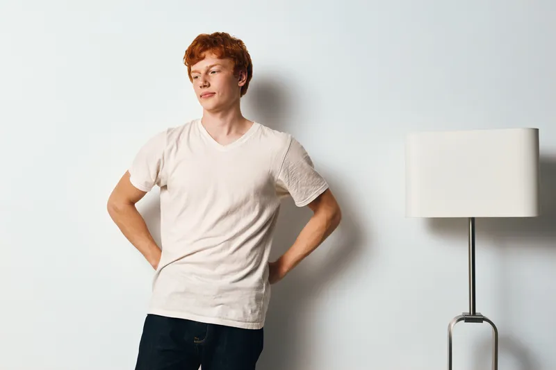 Portrait of young man with red hair standing near white wall and modern lamp wearing casual t shirt and jeans with hands on hips and thoughtful expression in minimal room.