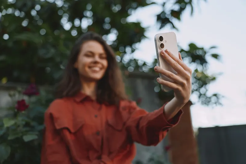 Young woman outdoors records a selfie in a rust shirt, smiling softly as she enjoys a casual lifestyle moment amid green foliage and a bright sky