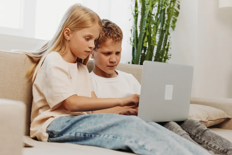 Two focused children using a laptop together on a sofa in a bright room with plants. Concept of learning, technology, cooperation, and modern childhood activities.