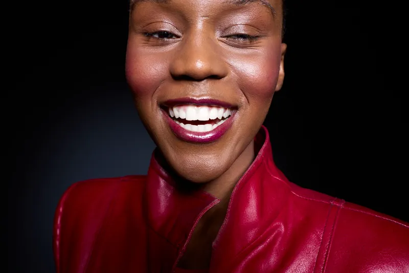 Close-up of happy African American woman with dark skin smiling with closed eyes wearing red leather jacket against black background. Concept of joy, fashion, beauty, and confidence.