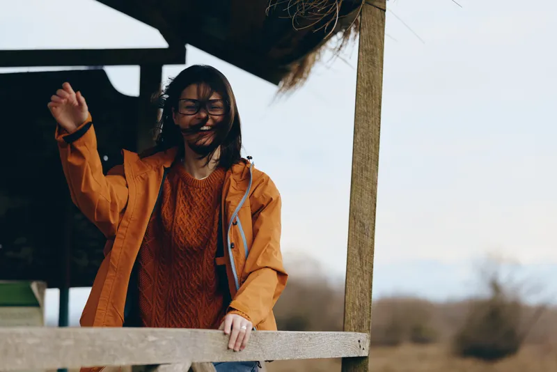 Smiling woman in orange jacket and glasses stands by wood railing outdoors, hand raised near a porch, lifestyle moment in cool brisk autumn light, natural daylight, quiet calm background