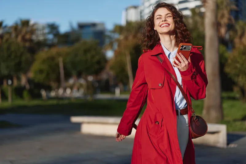 A smiling woman in a red coat walks in a sunny park, holding a smartphone. A lifestyle portrait capturing confidence, fashion, and outdoor leisure moments.