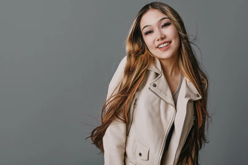 Young woman with long hair wearing beige jacket smiling confidently on gray background for portrait fashion beauty style casual look and happiness.
