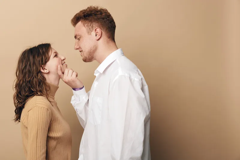 Young couple in casual clothes share intimate moment, man gently touches woman’s chin. Warm beige background creates cozy atmosphere. Concept of love, affection, and romantic connection.