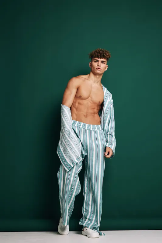Young man wearing striped oversized pants and shirt partially off shoulder poses against dark green studio background with confident expression and curly hair style.