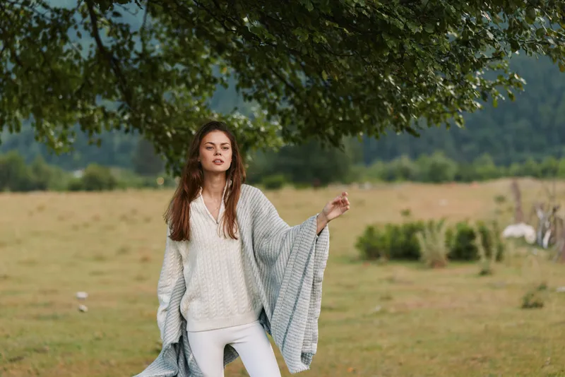 Young woman in a white knit sweater and shawl enjoys an outdoor lifestyle in a sunlit field, calm mood, natural textures, casual elegance, serene atmosphere