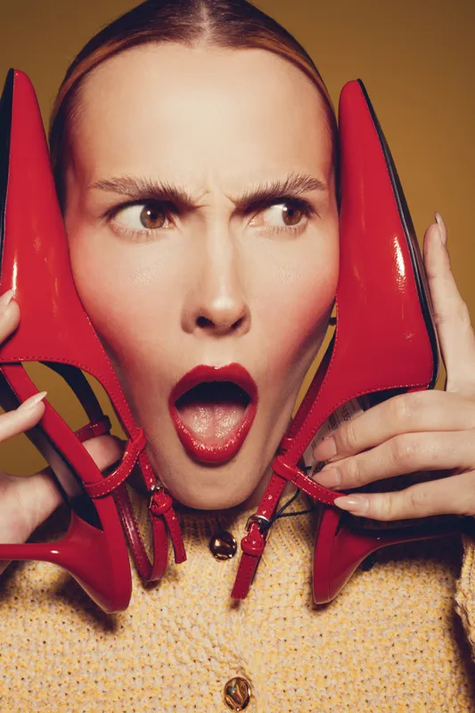 Surprised woman with red lipstick holds red high heel shoes near face, wearing yellow sweater. Concept of fashion, style, beauty, and shopping excitement in studio lighting.