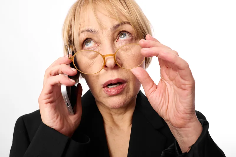 Senior woman holding phone to ear, adjusting glasses, surprised concerned expression, on white background, concept for communication stress, aging vision