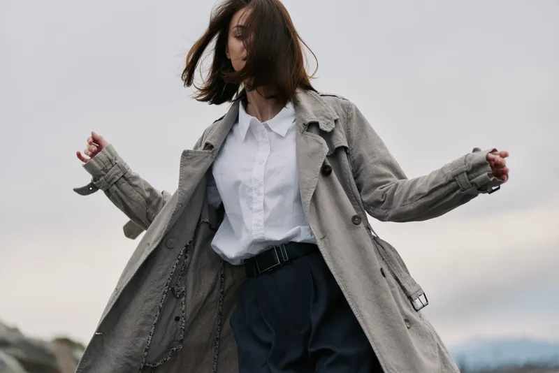 Young woman wearing long trench coat and white shirt poses outdoors with wind blowing through her hair. Casual fashion style in neutral tones against cloudy sky background.