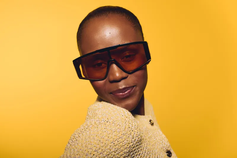 Confident black woman with dark skin wearing large sunglasses and a beige knitted sweater poses against yellow background. Concept of fashion, style, and modern beauty.