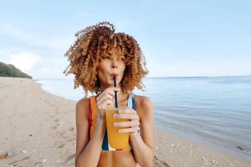 A young woman in a bikini on a sunny beach sips a cold juice through a straw, enjoying a refreshing moment. A candid lifestyle scene conveying summer leisure.