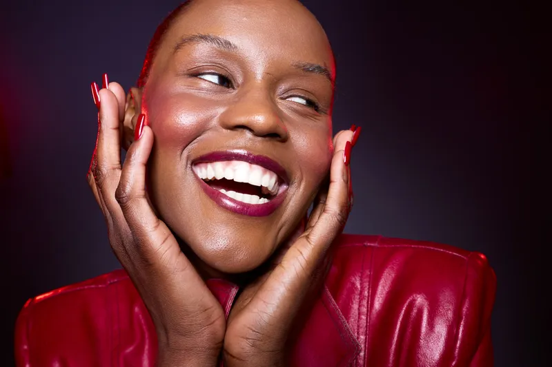 Close-up of a happy black woman with dark skin wearing a red leather jacket smiling broadly. She holds her face with manicured red nails against a dark studio background, beauty concept.