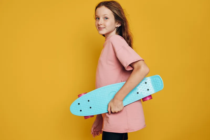 Young girl in a pink dress holding blue skateboard standing against yellow background looking confident.