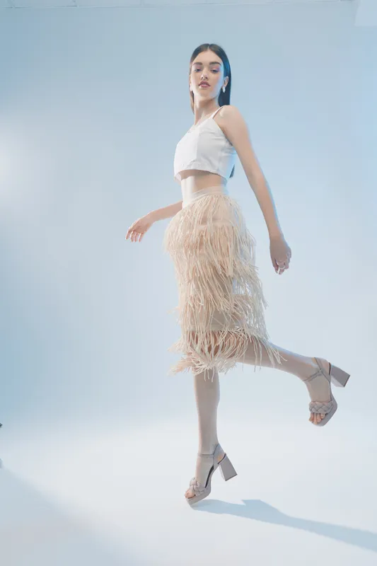 Young woman in a white crop top and beige fringed skirt poses on one leg against a light blue background. Concept of woman beauty fashion and elegance.