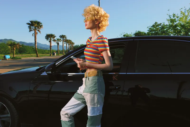 Cheerful woman outdoors, wearing striped top and patchwork jeans, leaning by a dark car, sunlit scene with palm trees, relaxed lifestyle and retro color mood