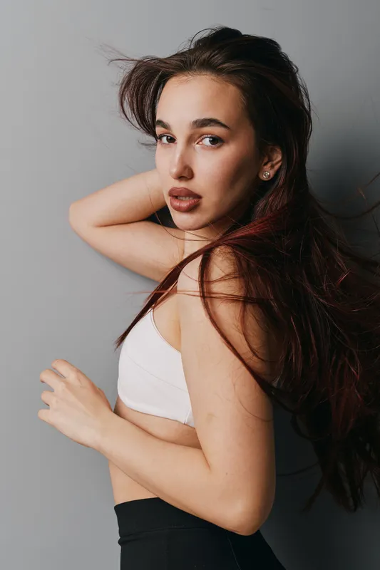 Young woman with long hair posing in white top and black pants against gray background. Beauty portrait with natural makeup and confident expression, studio shot with soft lighting.