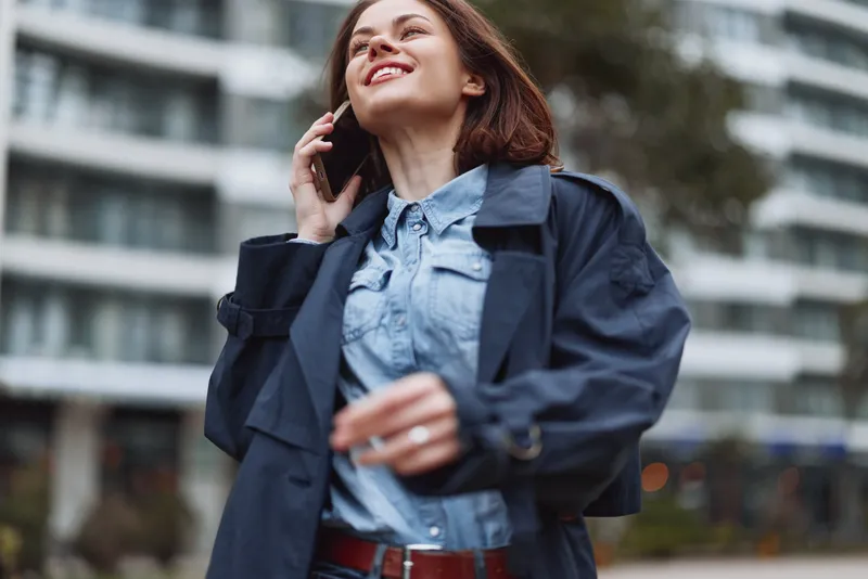 Young woman in a denim shirt and dark coat talks on her phone on a city street, smiling as she walks, with modern buildings and motionly urban backdrop nearby