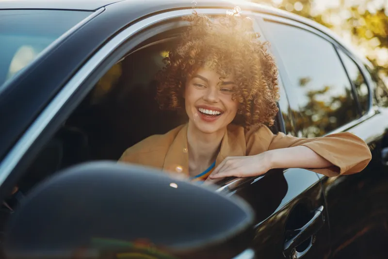 Smiling woman with curly hair in a car, enjoying a sunny day, casual lifestyle shot that highlights joy, freedom, and self expression, presented with warm retro film color and soft lighting.