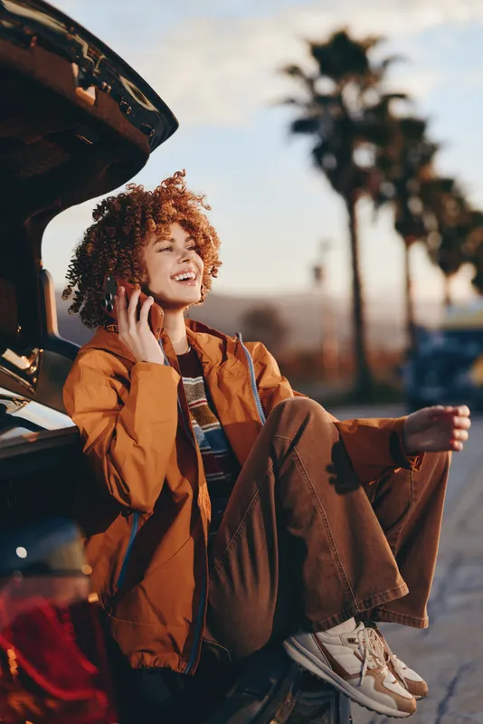 Woman lifestyle travel car on street outdoors at sunset, curly haired young woman in brown jacket holds phone near open trunk, palm trees blurred, warm golden light