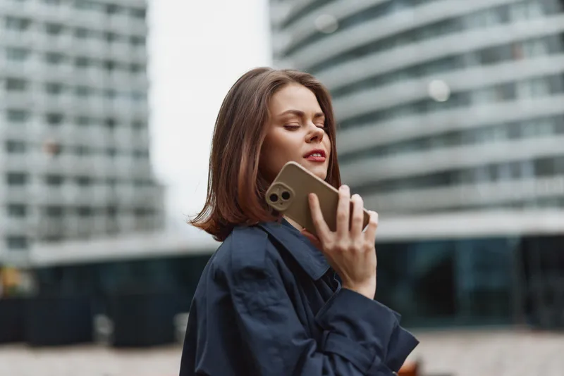 A woman with shoulder length hair stands outdoors in an urban setting, talking on a smartphone. She wears a dark coat and gazes away, conveying concentration and a moment of everyday communication in
