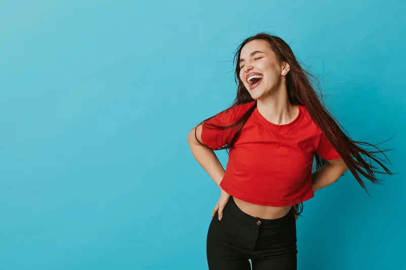 Young woman laughing with eyes closed standing against blue background wearing red shirt and black pants with hand on hip and long hair flowing in studio.