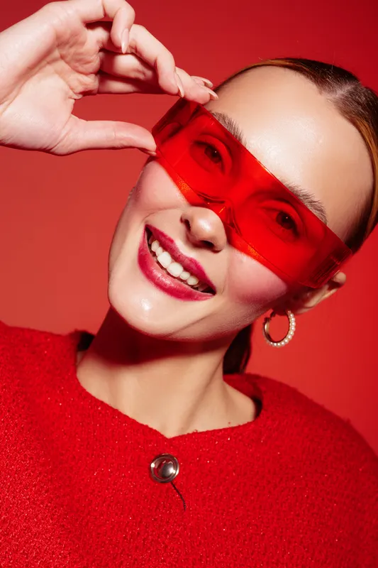Smiling woman with red sunglasses and red sweater poses against red background. Close-up portrait with hoop earrings and bright makeup. Concept of fashion, style, and confidence.
