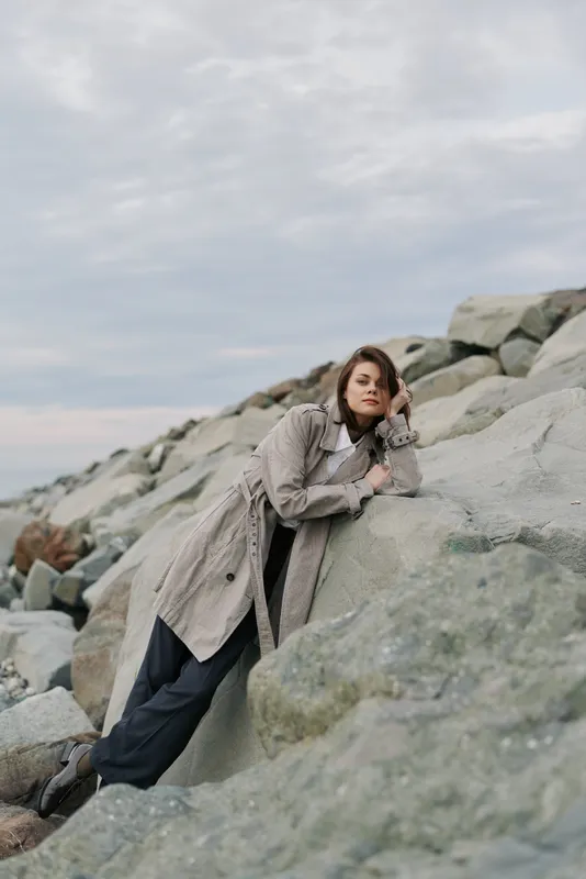 Young woman in cozy sweater and dark pants leans on large rocks outdoors under cloudy sky. Casual style and calm mood captured in natural rocky environment.