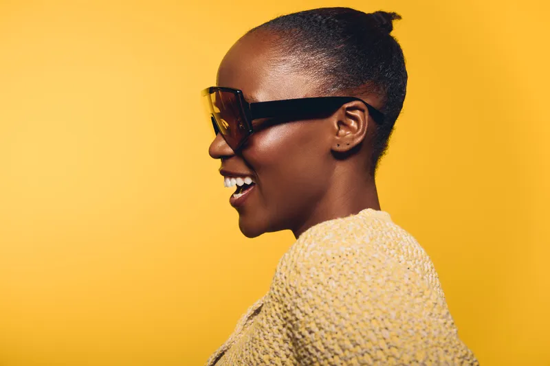 Smiling African woman with dark skin wearing large sunglasses and a knitted sweater against yellow background. Profile portrait captures joy, fashion, and confident mood with bright studio lighting.
