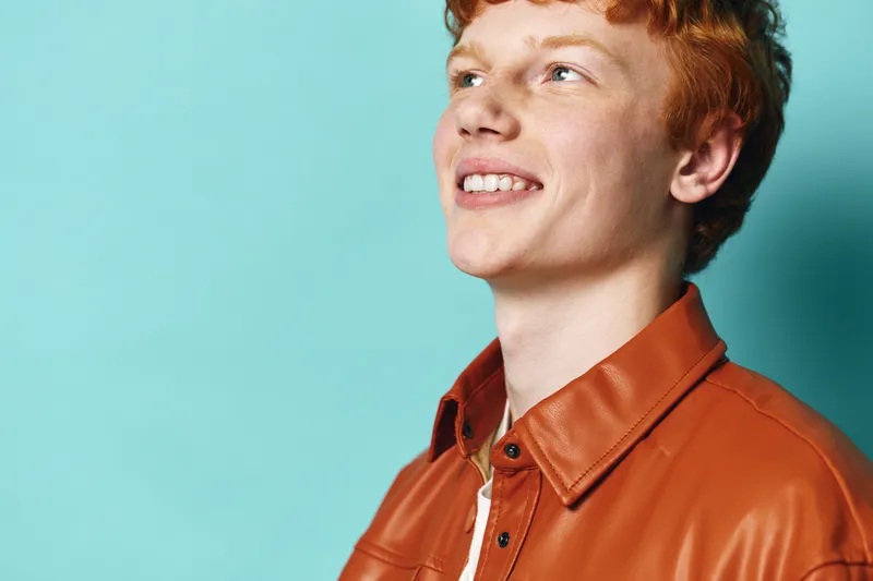 Portrait of smiling young man with red hair wearing brown leather jacket looking up against blue background. Casual style, positive expression, youth and confidence in studio shot.