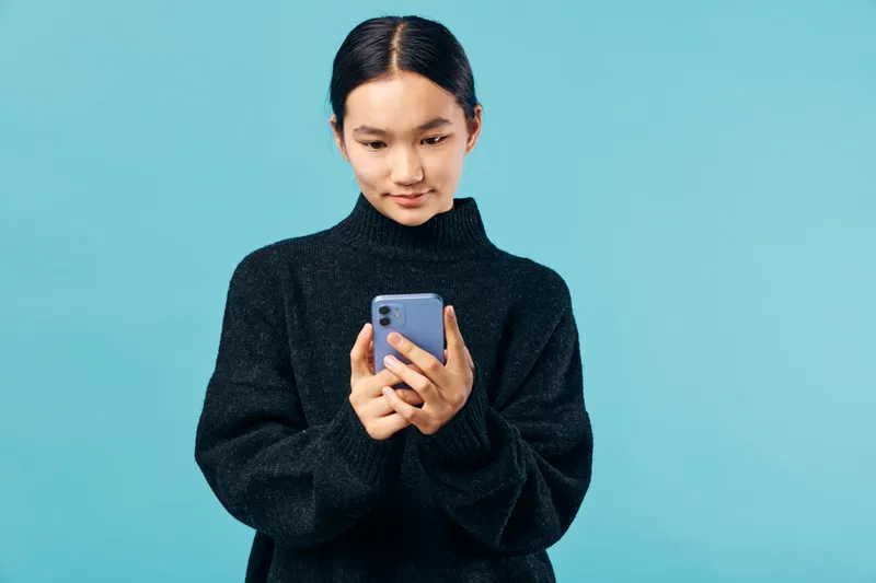 Young woman using smartphone with focused expression in casual black sweater on blue background. Female person holding mobile device for texting and online communication, digital technology and
