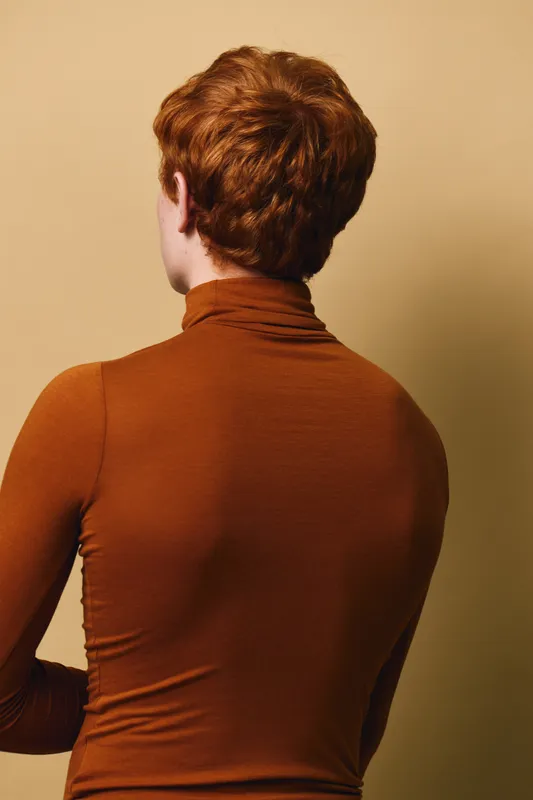 Person with short red hair wearing brown turtleneck top facing away with beige background. Studio portrait showing back and hairstyle with simple warm tones.