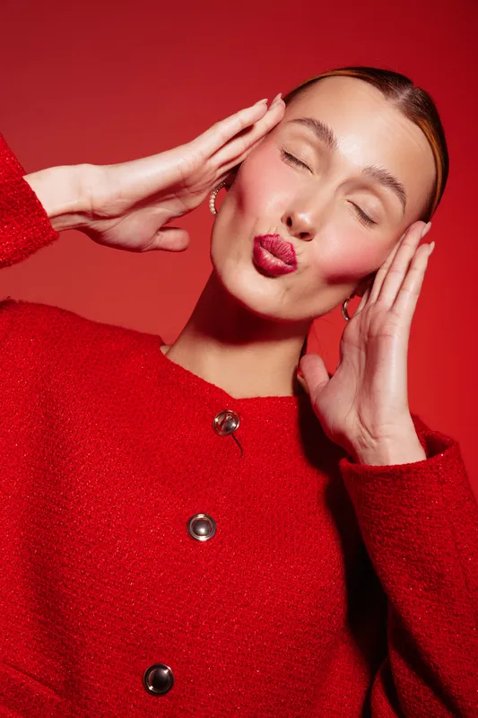 Young woman in red sweater makes kiss face with closed eyes and hands near head. Studio portrait on red background. Concept of beauty, fashion, emotion, and playful mood.
