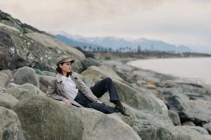 A thoughtful woman in casual outfit and cap relaxes on large rocks by the seaside with mountains in the background. Concept of outdoor leisure and nature escape.