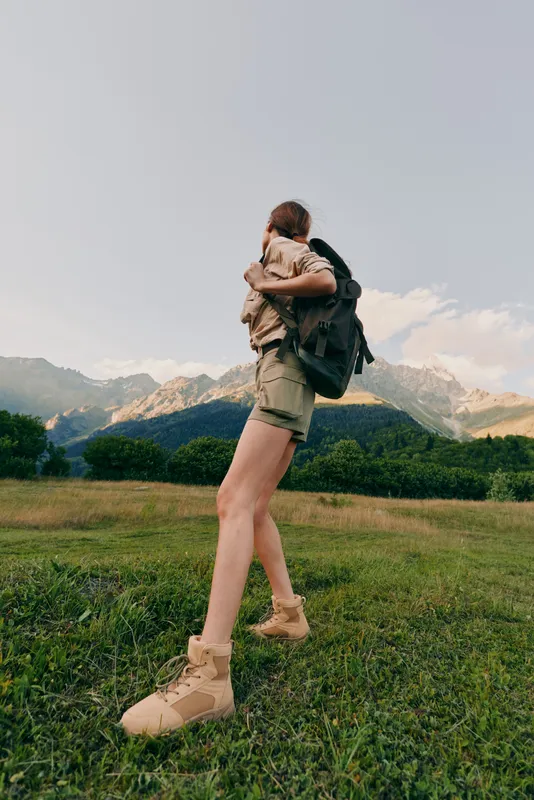 Woman hiking outdoors with a child on her back, backpack and boots in a meadow beneath mountains, nature, adventure, and family lifestyle.