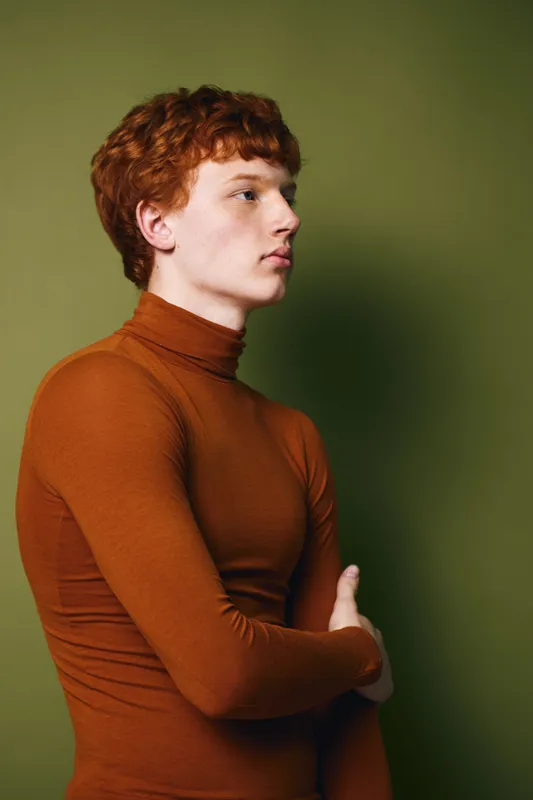 Young man with red hair and pale skin wearing brown turtleneck standing in profile against green background looking serious and thoughtful with arms crossed.