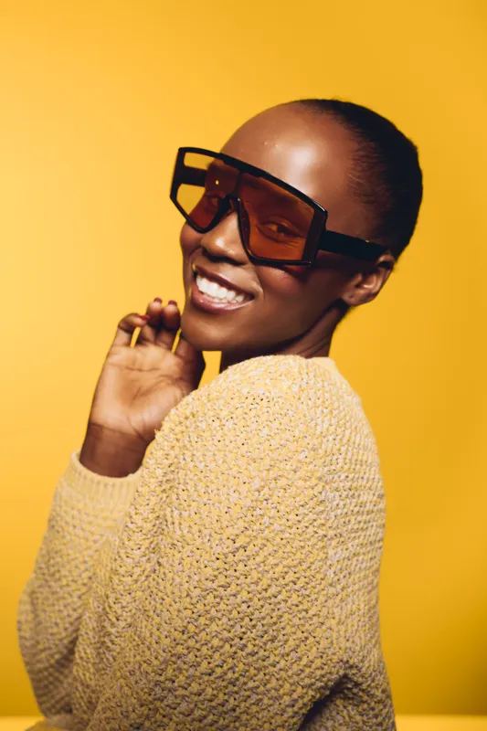 Smiling African American woman with dark skin wearing oversized sunglasses and a knitted sweater poses against a bright yellow background. Concept for fashion, style, and confidence.
